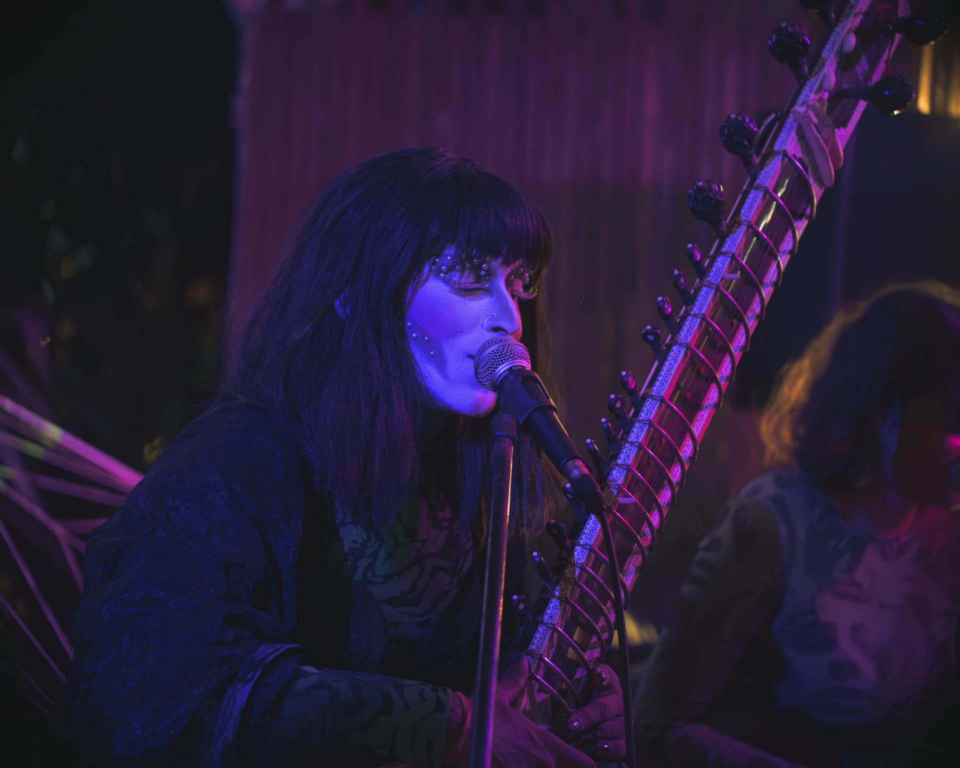 Vocalist singing beside a sitar in deep blue and magenta light